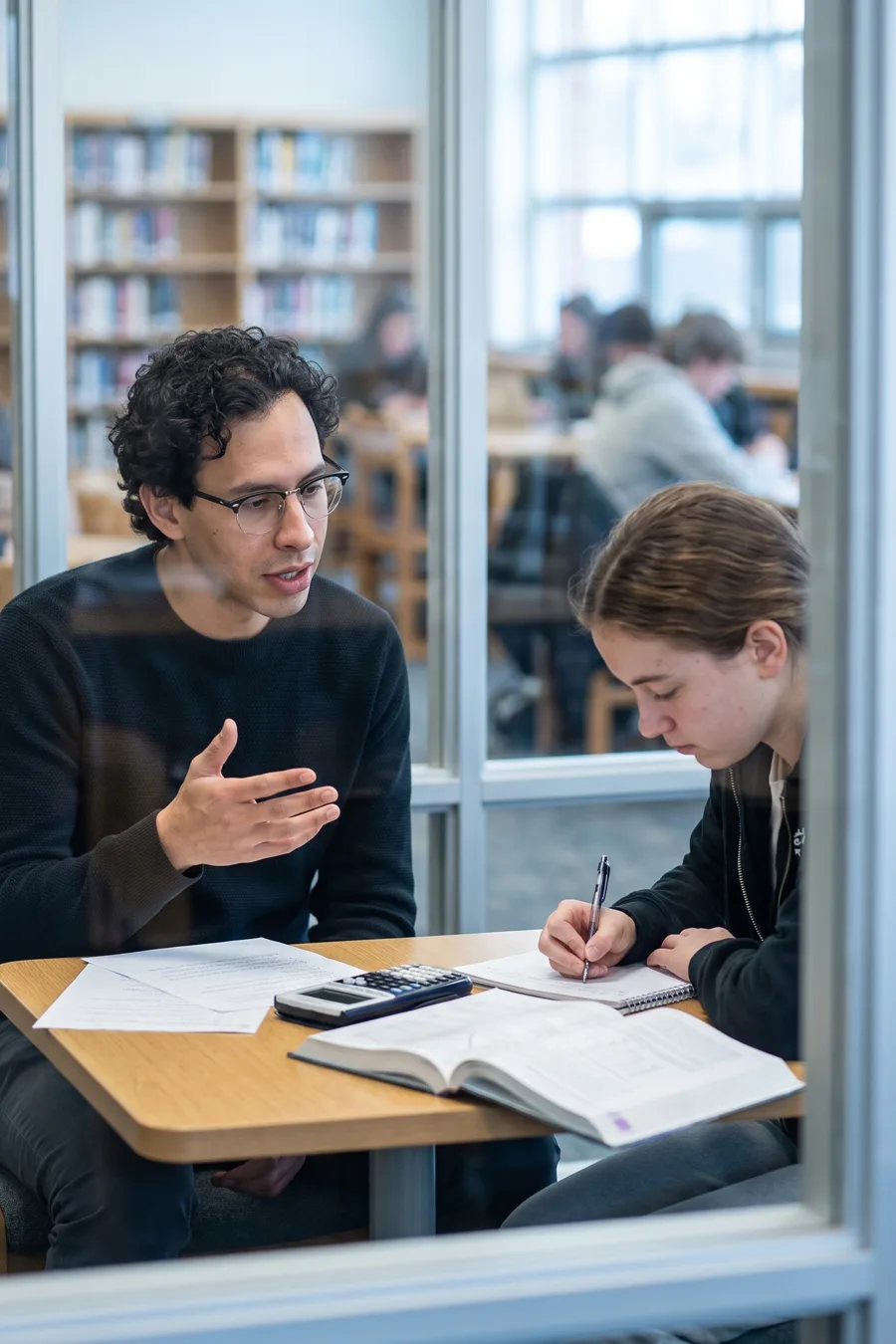 Nico talking a student through a problem in a glass-walled library study room